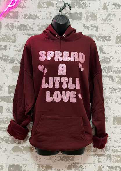 a maroon sweatshirt that says "Spread A Little Love" in a pink glitter graphic with hearts.