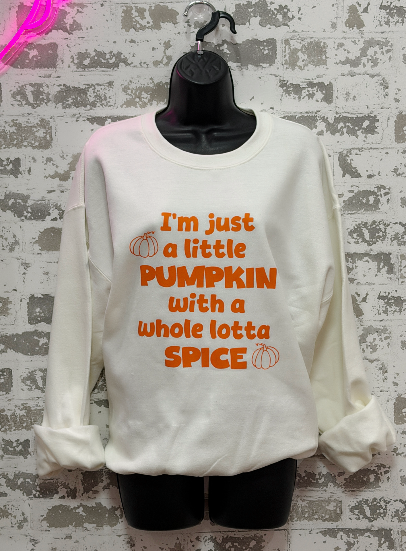 An off-white crewneck sweatshirt with orange text reading 'I'm just a little PUMPKIN with a whole lotta SPICE' and small pumpkin graphics, displayed on a black hanger/mannequin against a faux brick wall.