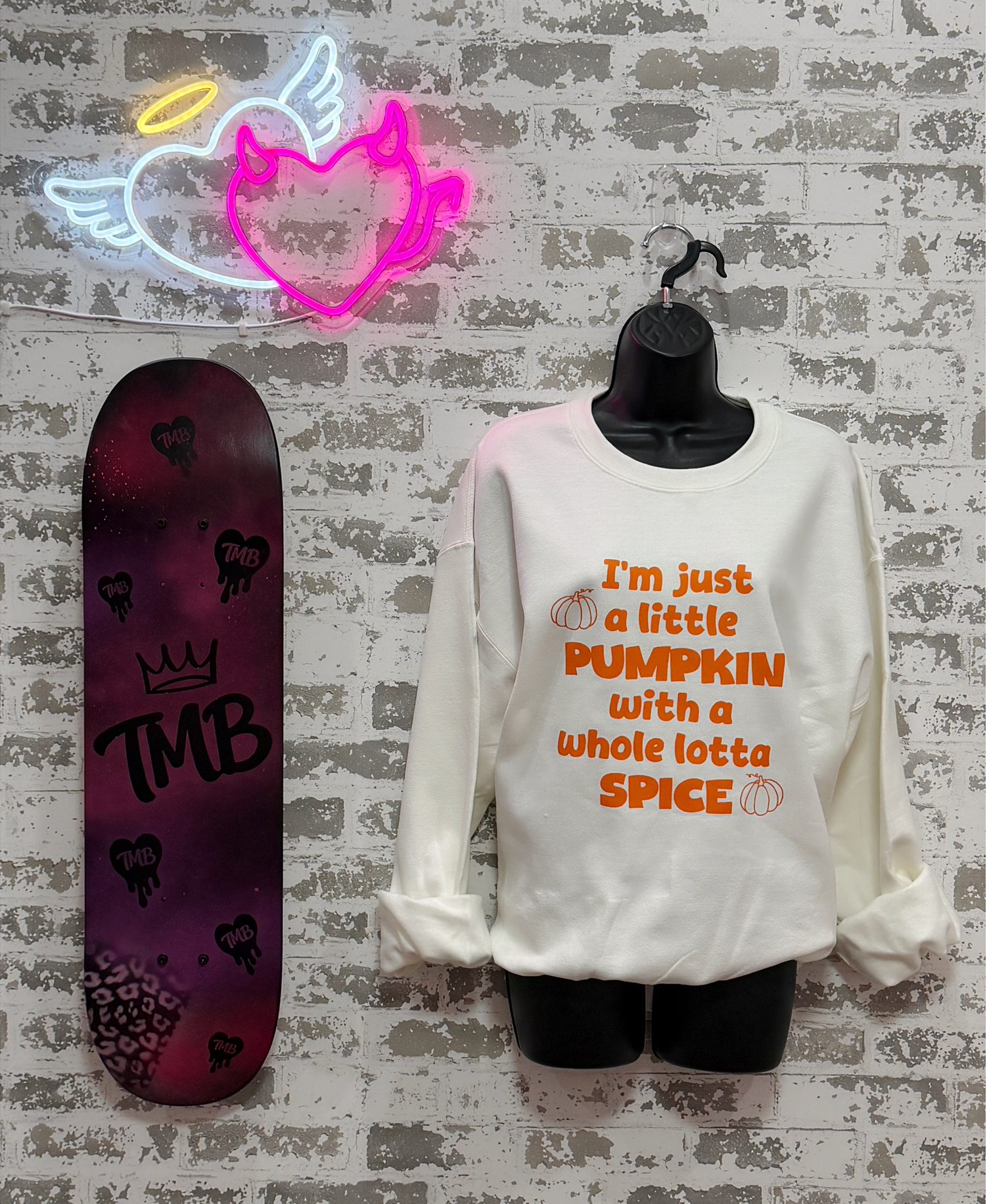 A white long-sleeve sweatshirt with orange text reading 'I'm just a little PUMPKIN with a whole lotta SPICE' is displayed on a black mannequin torso against a white brick wall. A neon sign hangs above, and a skateboard stands to the left.