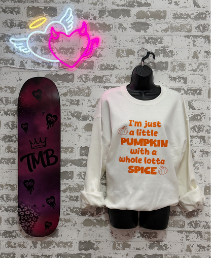 A white long-sleeve sweatshirt with orange text reading 'I'm just a little PUMPKIN with a whole lotta SPICE' is displayed on a black mannequin torso against a white brick wall. A neon sign hangs above, and a skateboard stands to the left.