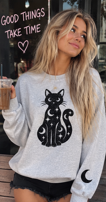 A woman wearing a light gray crewneck sweatshirt with a black cat and moon graphic, holding an iced coffee in front of a storefront window.