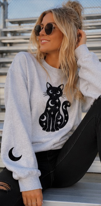 A woman wearing a light gray crewneck sweatshirt with a black cat and moon graphic, paired with black jeans, sitting on outdoor bleachers.