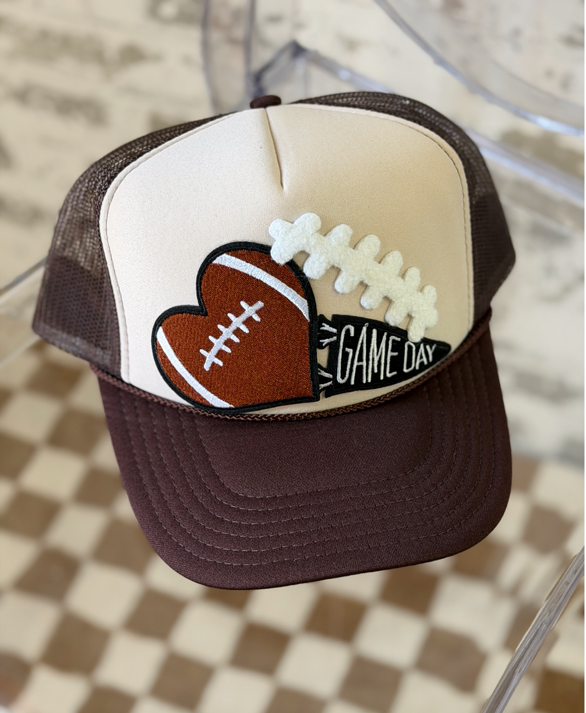 A woman wearing a cream and chocolate brown high-crown trucker hat featuring a dimensional football-heart patch and a GAME DAY pennant embroidery.