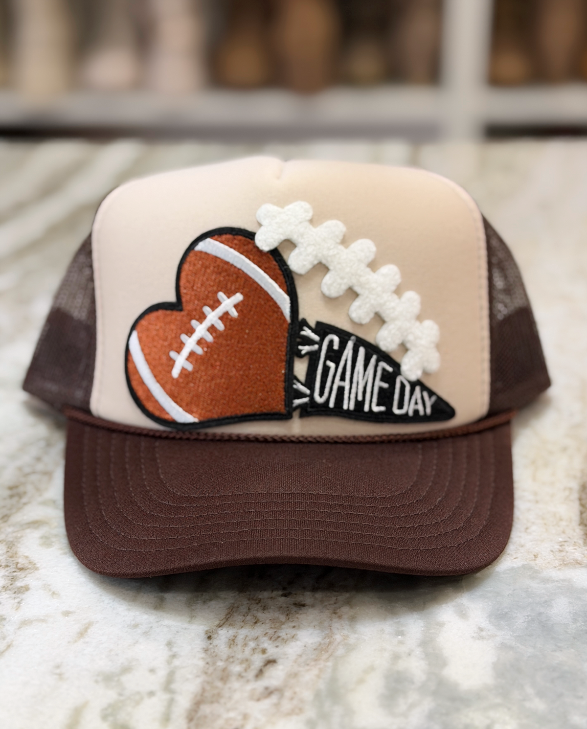 The Game Day Heart Trucker Hat features a cream front panel with a chocolate brown bill and mesh back, showcasing an embroidered football-shaped heart patch paired with a GAME DAY pennant detail.