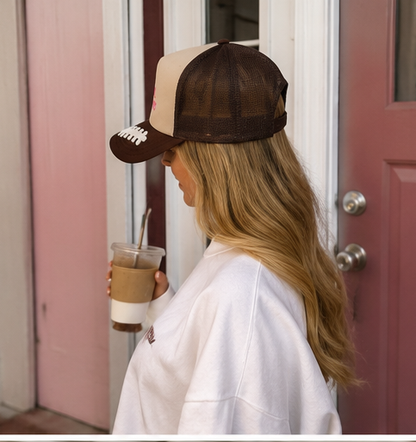 A woman in a white sweatshirt is shown from the side profile, wearing a brown and beige trucker-style baseball cap with white embroidered details, holding an iced coffee in front of a pink door.