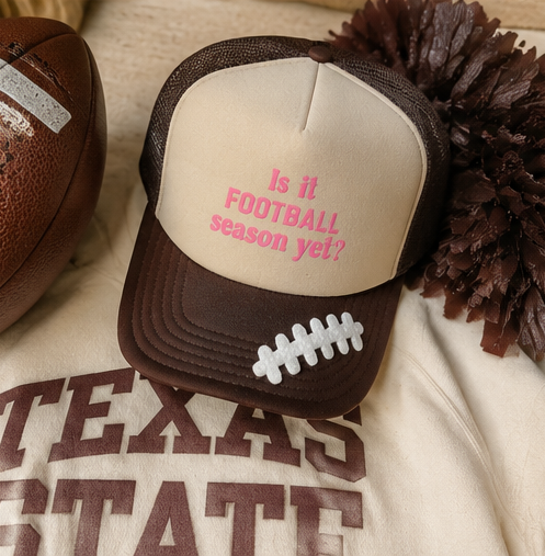 A brown and beige trucker hat with pink text reading 'Is it FOOTBALL season yet?' and a white laces graphic on the brim, styled on a Texas State sweatshirt with a football and pom-pom nearby.