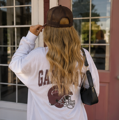 A woman viewed from behind wearing a white long-sleeve sweatshirt with a brown football helmet graphic and 'GAMEDAY' text, a brown mesh-back cap, and carrying a black shoulder bag, standing in front of a pink door and window.