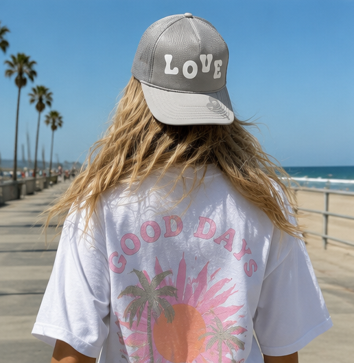 A person standing on a beach boardwalk wearing a gray baseball cap with the word 'LOVE' in white letters on the front and a white graphic T-shirt with pink text and a palm tree sun design. The background shows a sunny beach, ocean, and palm trees.