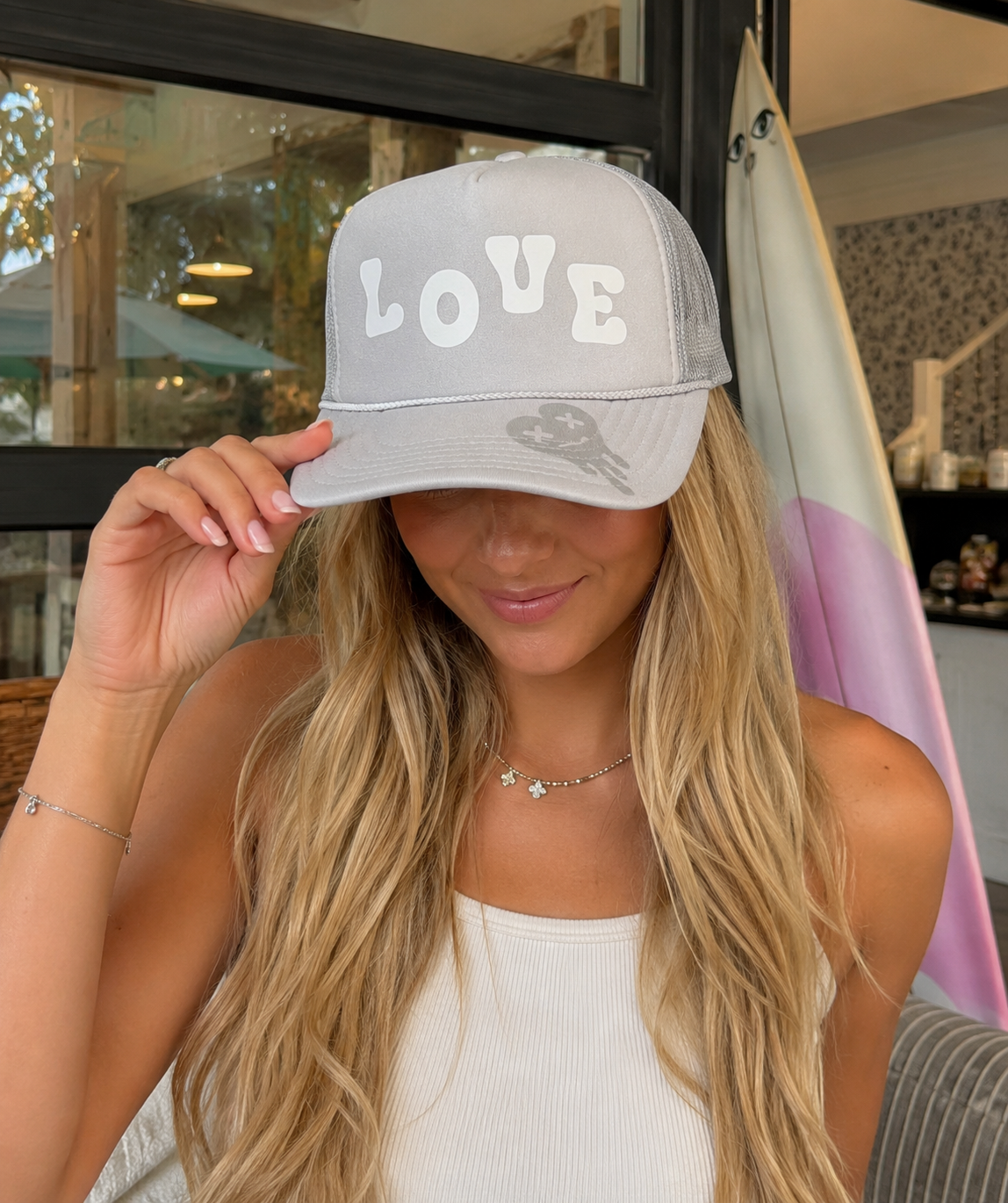 A woman wearing a light gray trucker-style baseball cap with the word 'LOVE' in white across the front, photographed indoors near a window and surfboard.