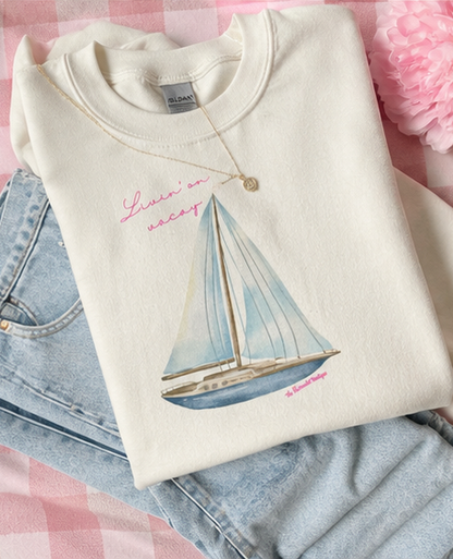 A folded cream sweatshirt with a sailboat graphic and pink script text, styled on top of light blue jeans against a pink gingham background with a pink flower.