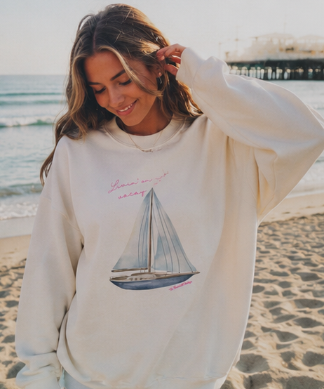 A woman on a sandy beach wearing a cream-colored sweatshirt with a sailboat graphic and pink script text, with the ocean and a pier in the background.