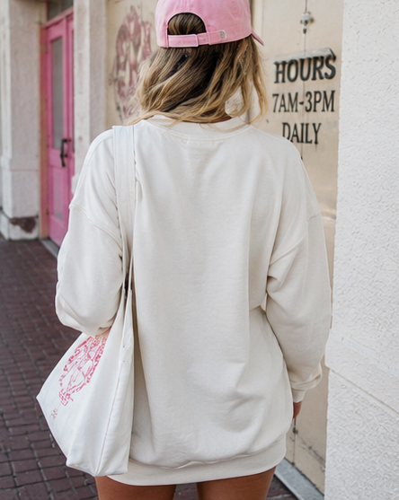 A woman viewed from behind wearing an oversized cream sweatshirt, a pink baseball cap, and carrying a white tote bag with a pink graphic print, standing outdoors against a building wall.