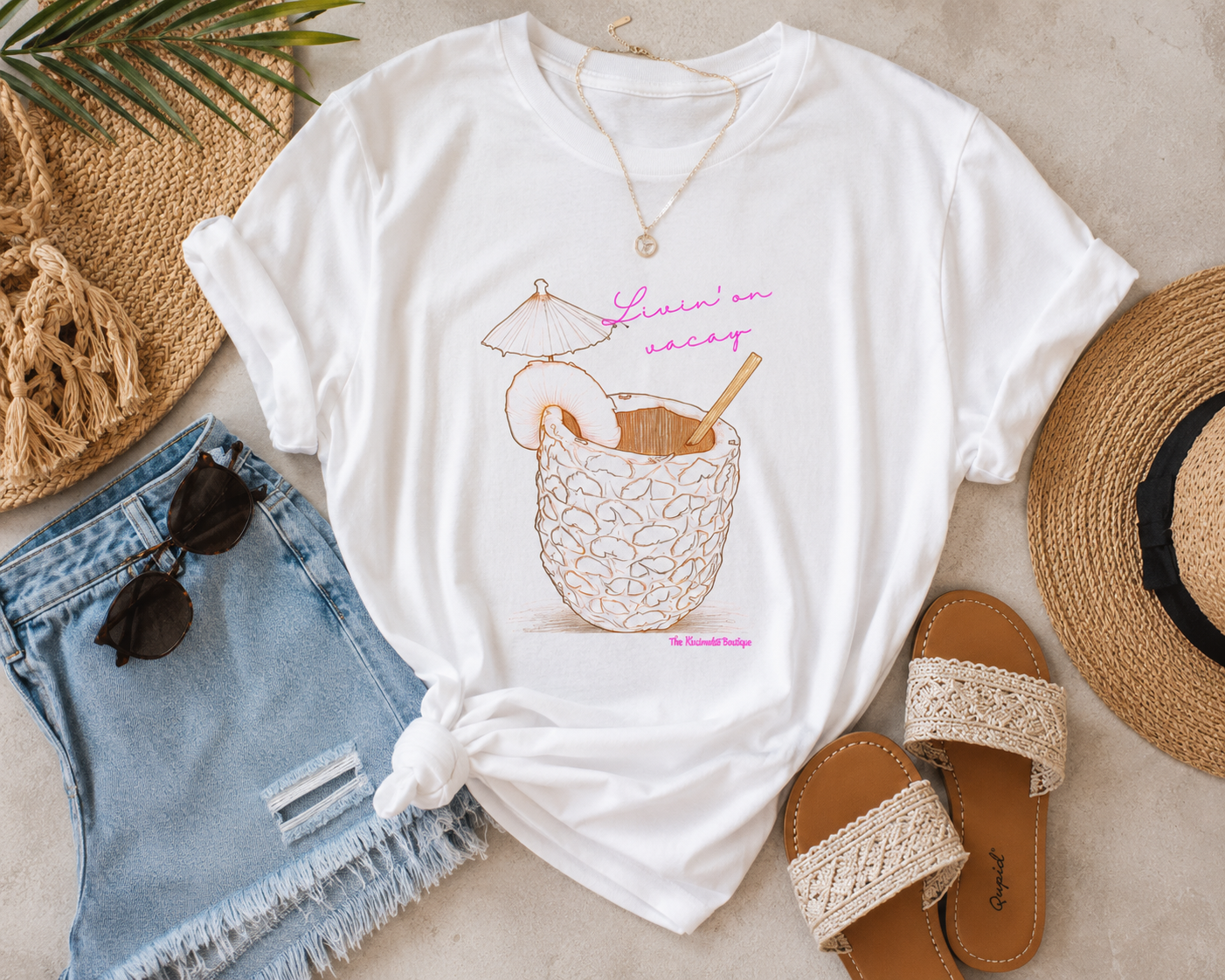 White t-shirt with a graphic design of a pineapple drink, sunglasses, denim shorts, and sandals on a neutral background.