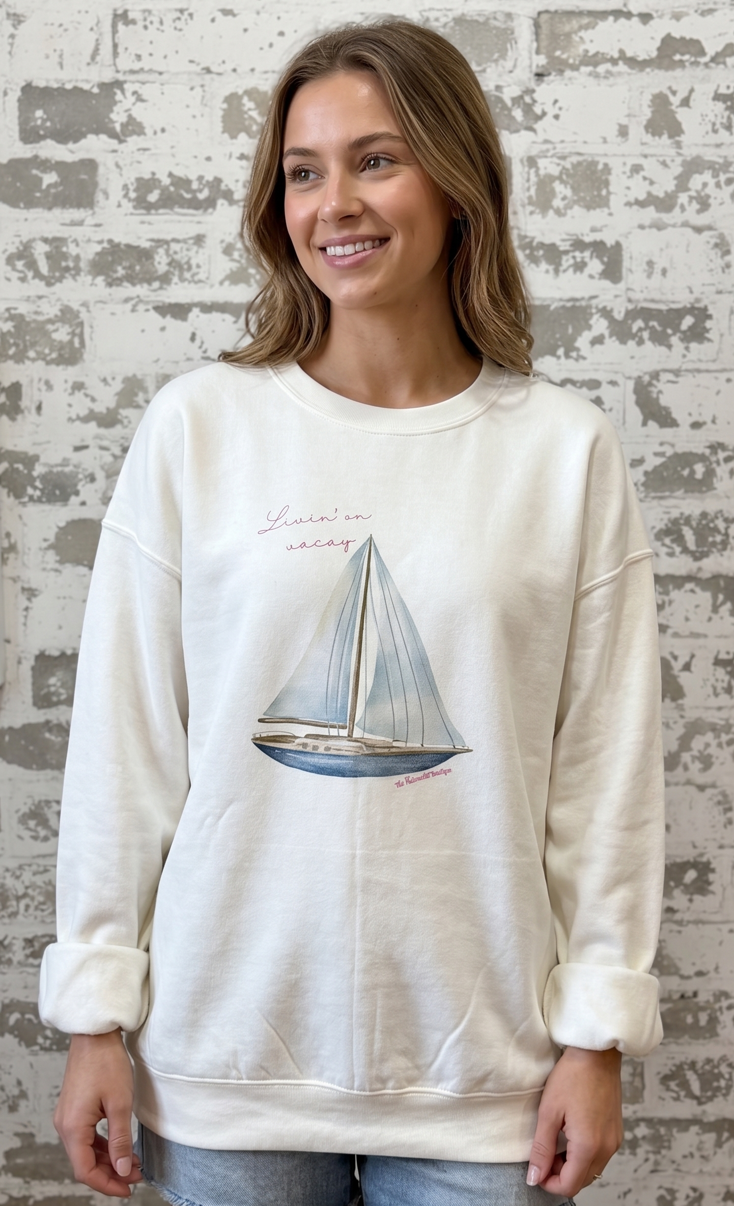 Woman wearing a cream sweatshirt with a sailboat design against a textured wall.