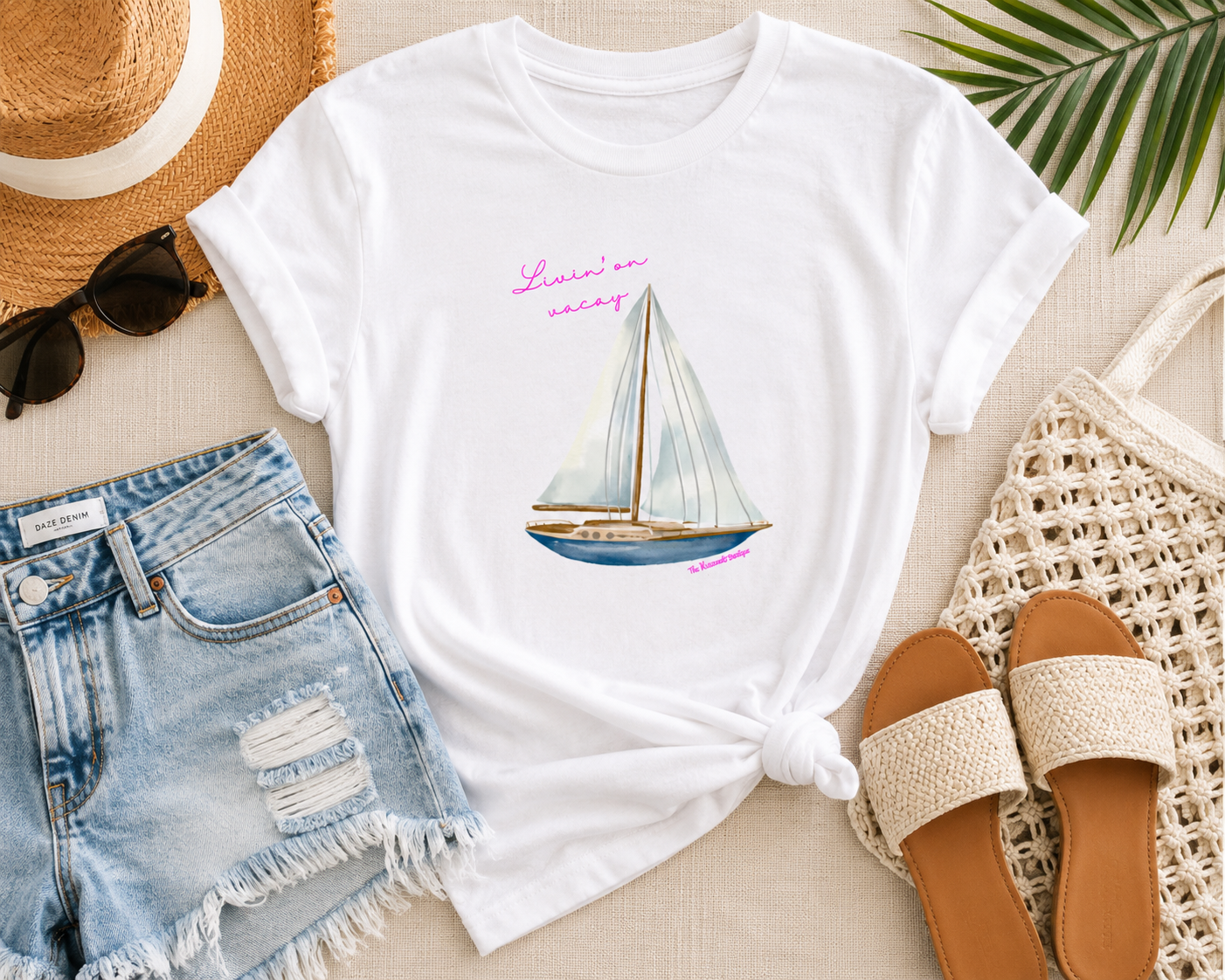 White t-shirt with sailboat design, denim shorts, straw hat, sunglasses, and sandals on a light surface.