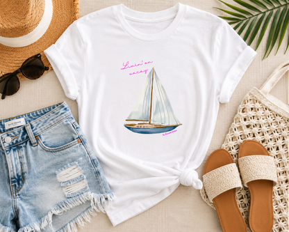White t-shirt with sailboat design, denim shorts, straw hat, sunglasses, and sandals on a light surface.