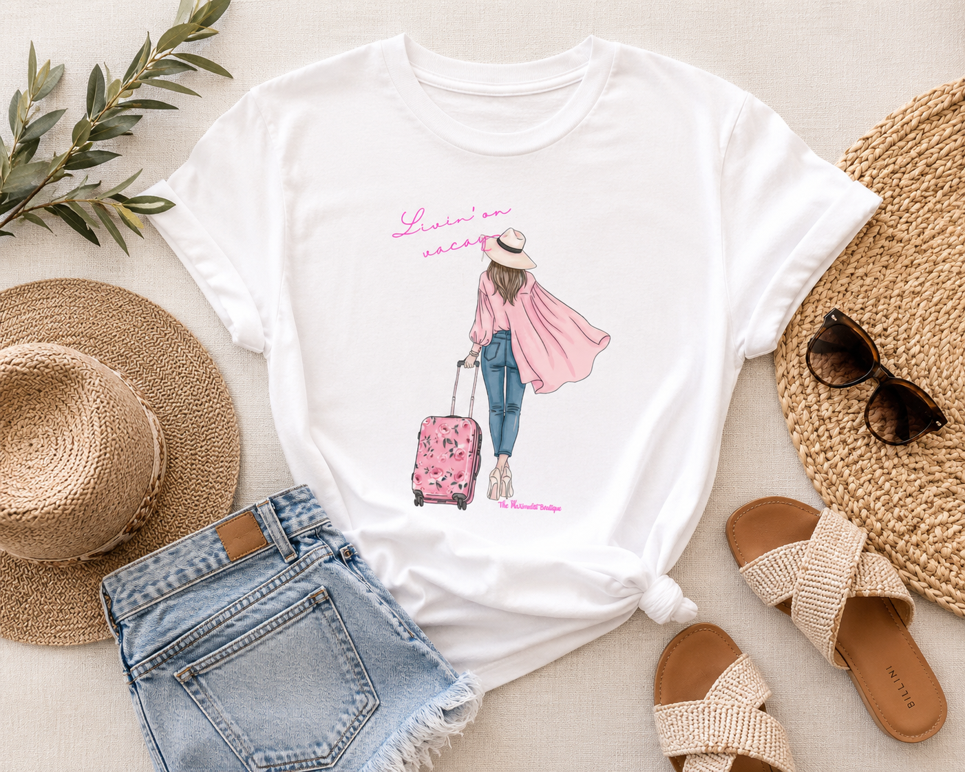 White t-shirt with a graphic design of a person with a suitcase, surrounded by a straw hat, sunglasses, sandals, and denim shorts on a light surface.