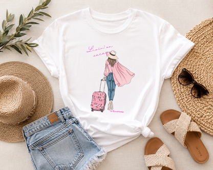 White t-shirt with a graphic design of a person with a suitcase, surrounded by a straw hat, sunglasses, sandals, and denim shorts on a light surface.