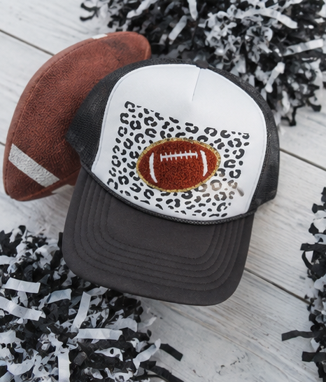 A football-themed trucker hat with a white front panel featuring a leopard print pattern and a central embroidered football, displayed on a white wood background with a plush football and black-and-white shredded paper accents.