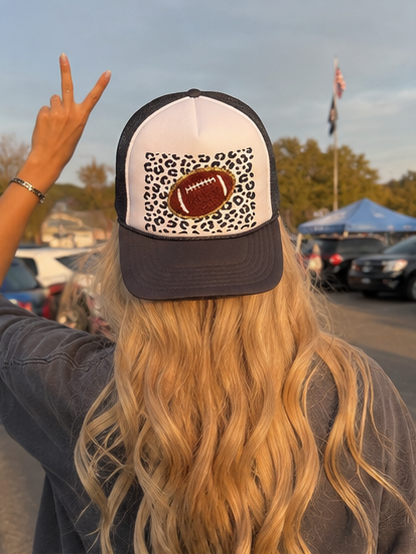 A woman viewed from behind wearing a trucker hat with a white front panel, dark gray brim and back, and a leopard-print patch featuring a football graphic. She is outdoors at what looks like a tailgate or parking lot with cars and tents in the background.
