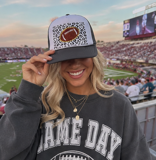 A smiling woman in a stadium crowd wearing a gray and white trucker hat with a leopard print and football graphic, along with a dark gray sweatshirt that says 'GAME DAY'.