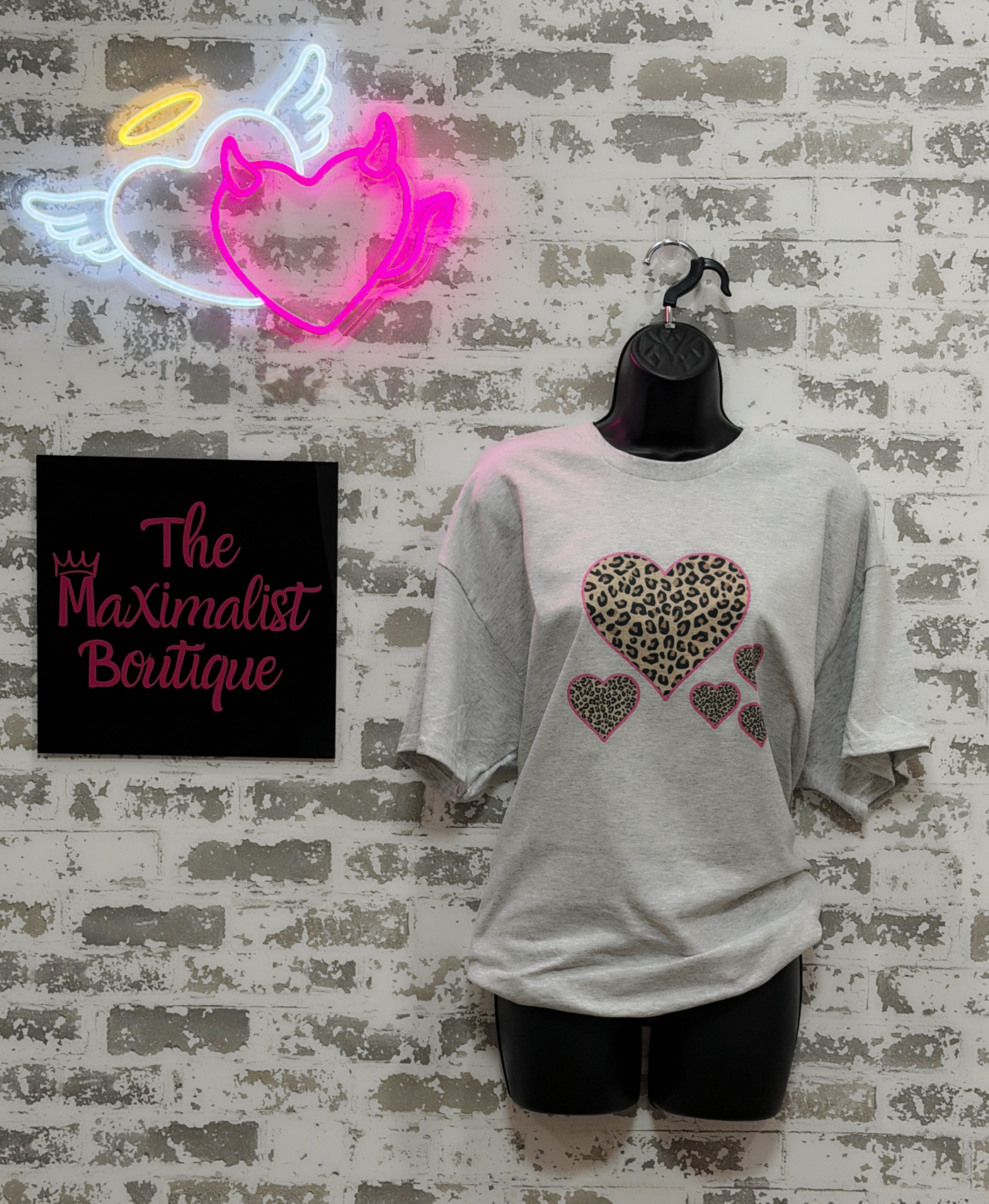 Mannequin wearing a gray t-shirt with heart designs in front of a neon sign and 'The Maximalist Boutique' sign.
