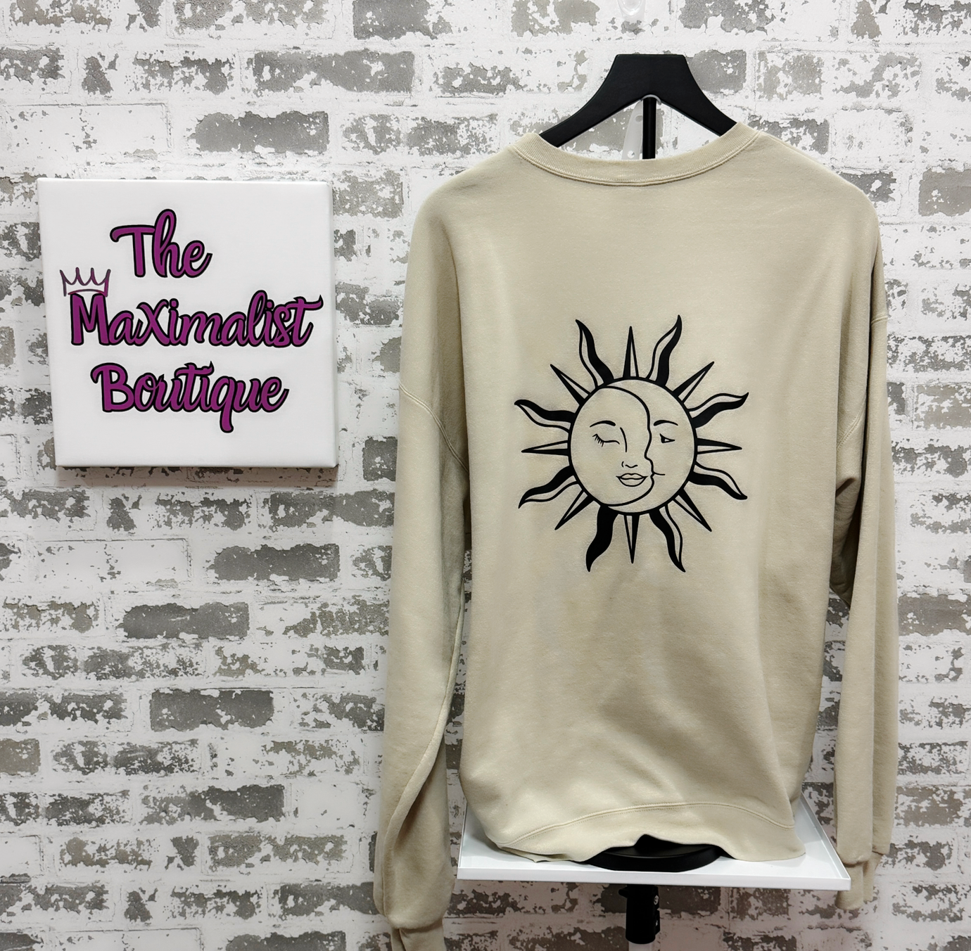Beige sweatshirt with a sun design on a hanger against a textured wall with 'The Maximalist Boutique' sign.
