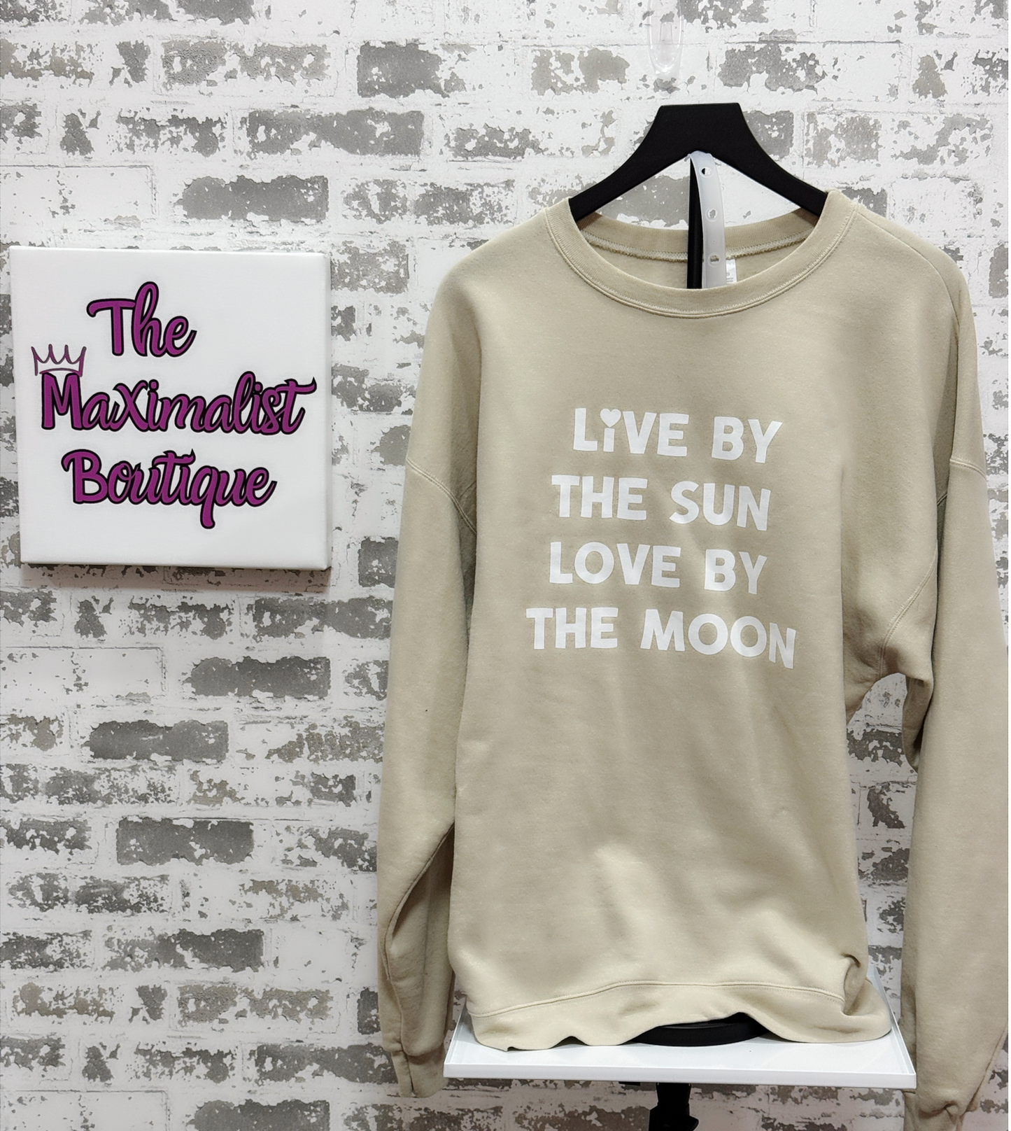 Beige sweatshirt with text 'Live by the sun, love by the moon' on a hanger against a brick wall with 'The Maximalist Boutique' sign.