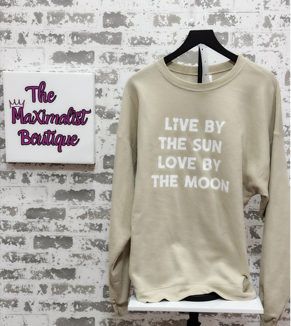 Beige sweatshirt with text 'Live by the sun, love by the moon' on a hanger against a brick wall with 'The Maximalist Boutique' sign.