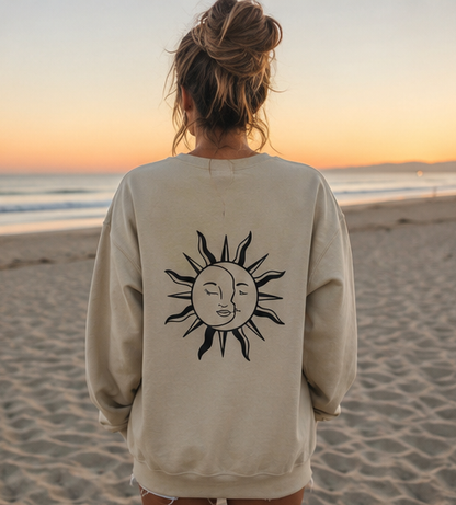 A woman stands on a sandy beach at sunset wearing an oversized cream sweatshirt with a black line-art sun graphic printed on the back.