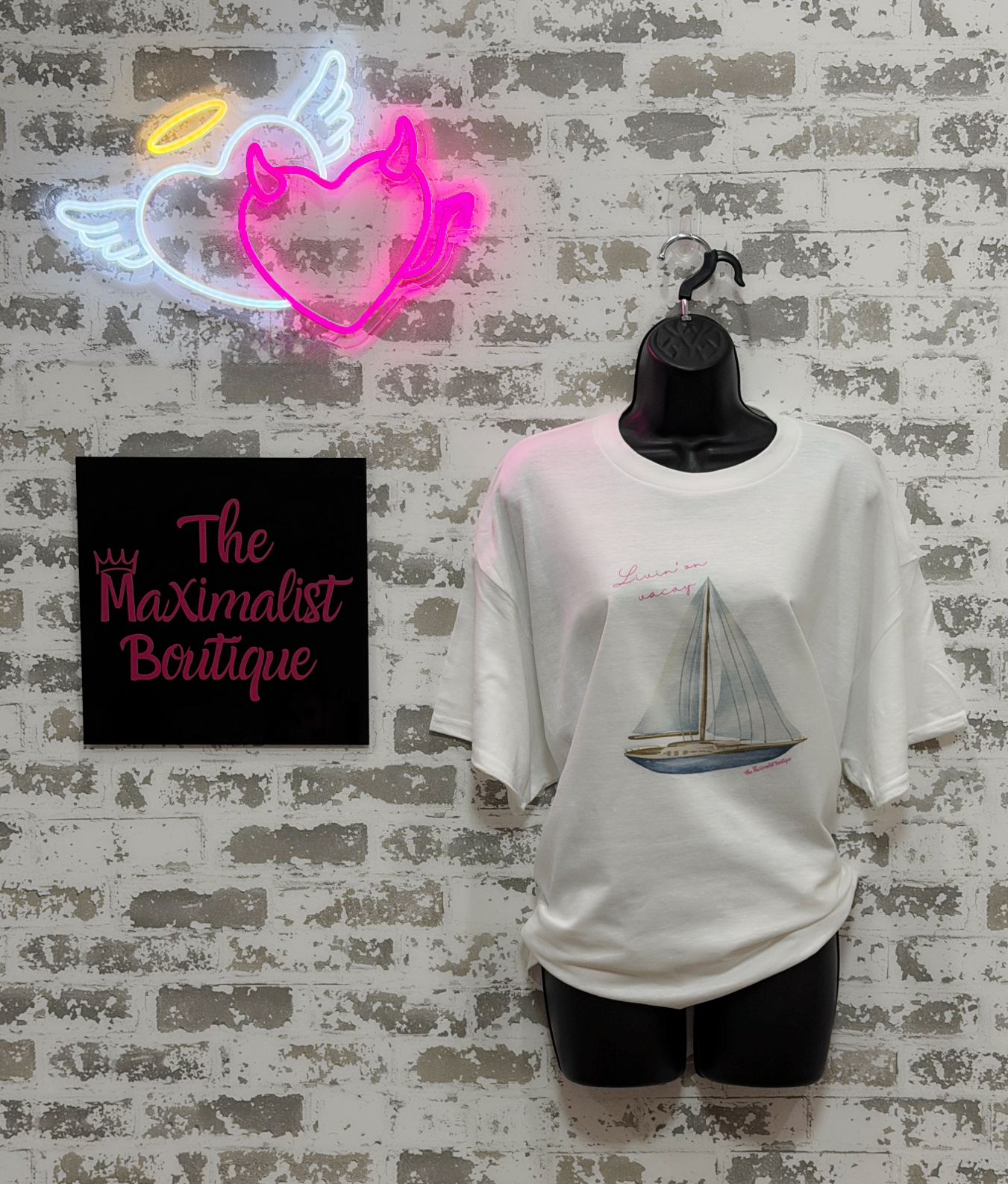 White t-shirt with sailboat design on a mannequin against a textured wall with neon sign and 'The Maximalist Boutique' sign.