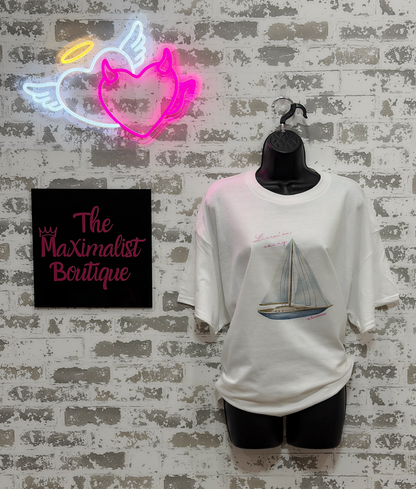 White t-shirt with sailboat design on a mannequin against a textured wall with neon sign and 'The Maximalist Boutique' sign.