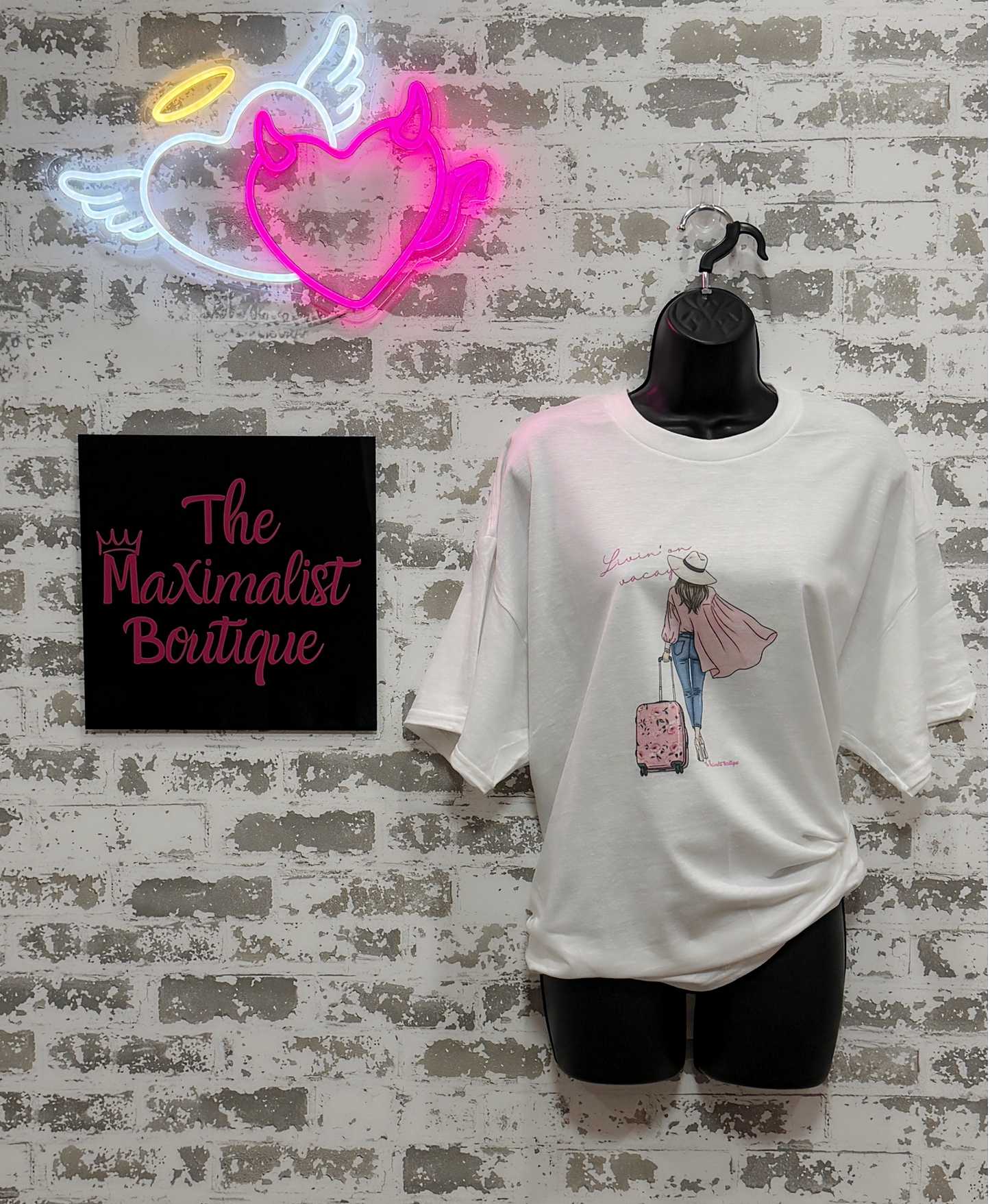 White t-shirt with graphic design on a mannequin against a textured wall with neon sign and 'The Maximalist Boutique' sign.