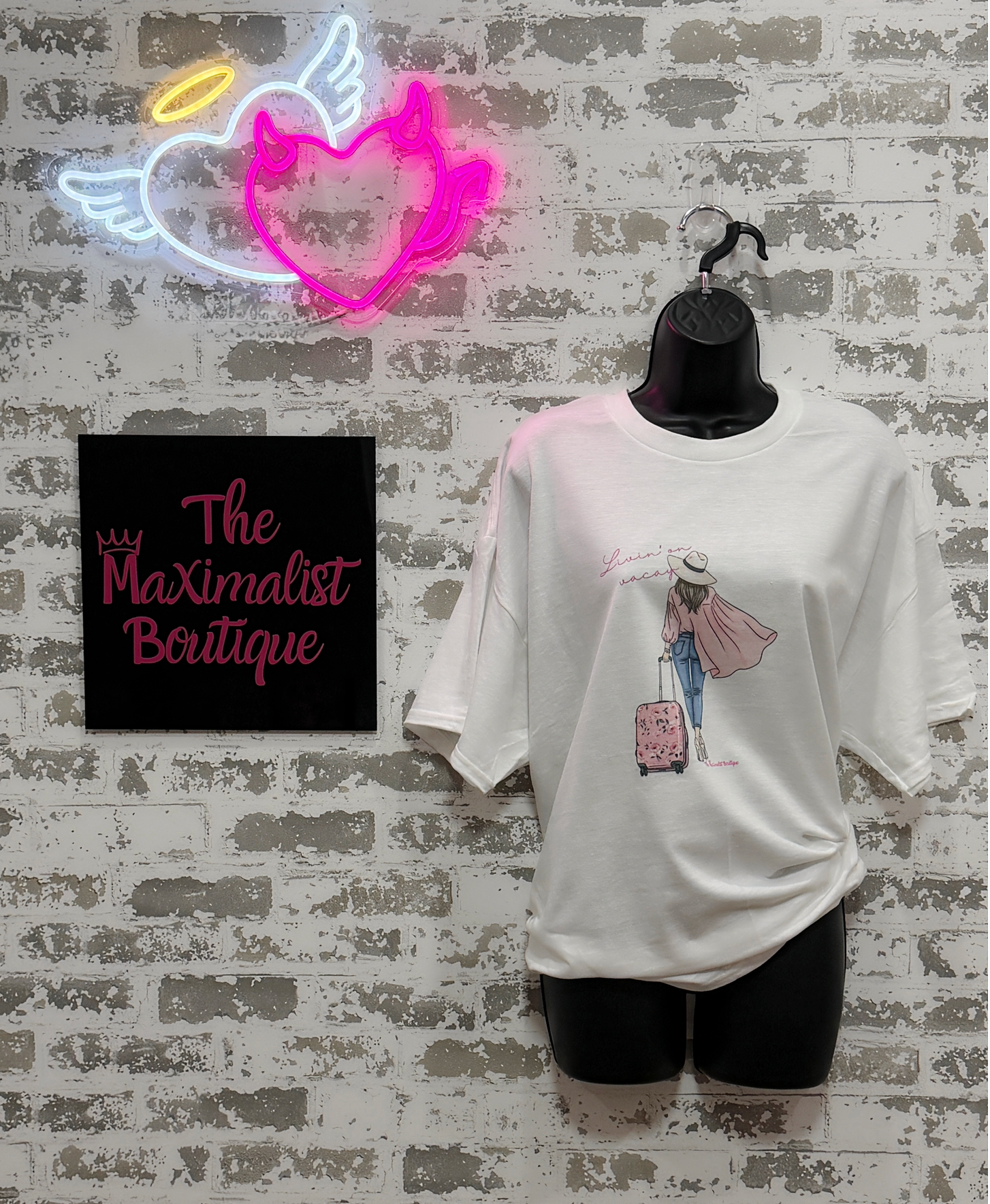 White t-shirt with graphic design on a mannequin against a textured wall with neon sign and 'The Maximalist Boutique' sign.