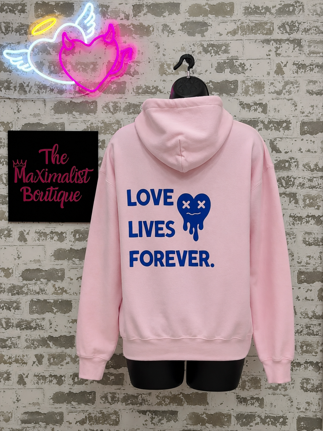 Pink hoodie with 'LOVE LIVES FOREVER' text on a textured wall background with neon sign.