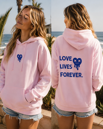 Woman wearing a pink hoodie with 'Love Lives Forever' text and blue heart design, standing by the ocean.