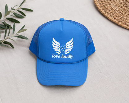 Blue cap with 'love loudly' text and logo on a light gray surface with a plant and woven mat.