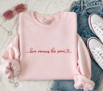 A folded light pink sweatshirt with the phrase 'love remains the same' in red cursive script across the chest, styled in a flat lay with blue jeans, white sneakers, sunglasses, and jewelry on a white textured background.