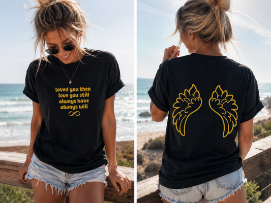 Woman wearing a black t-shirt with gold text and design, standing by the ocean.