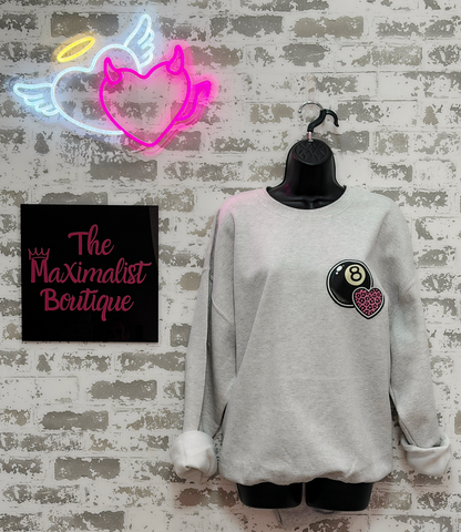 Gray sweatshirt with a heart and number 8 design on a hanger against a textured wall with a neon sign and 'The Maximalist Boutique' sign.