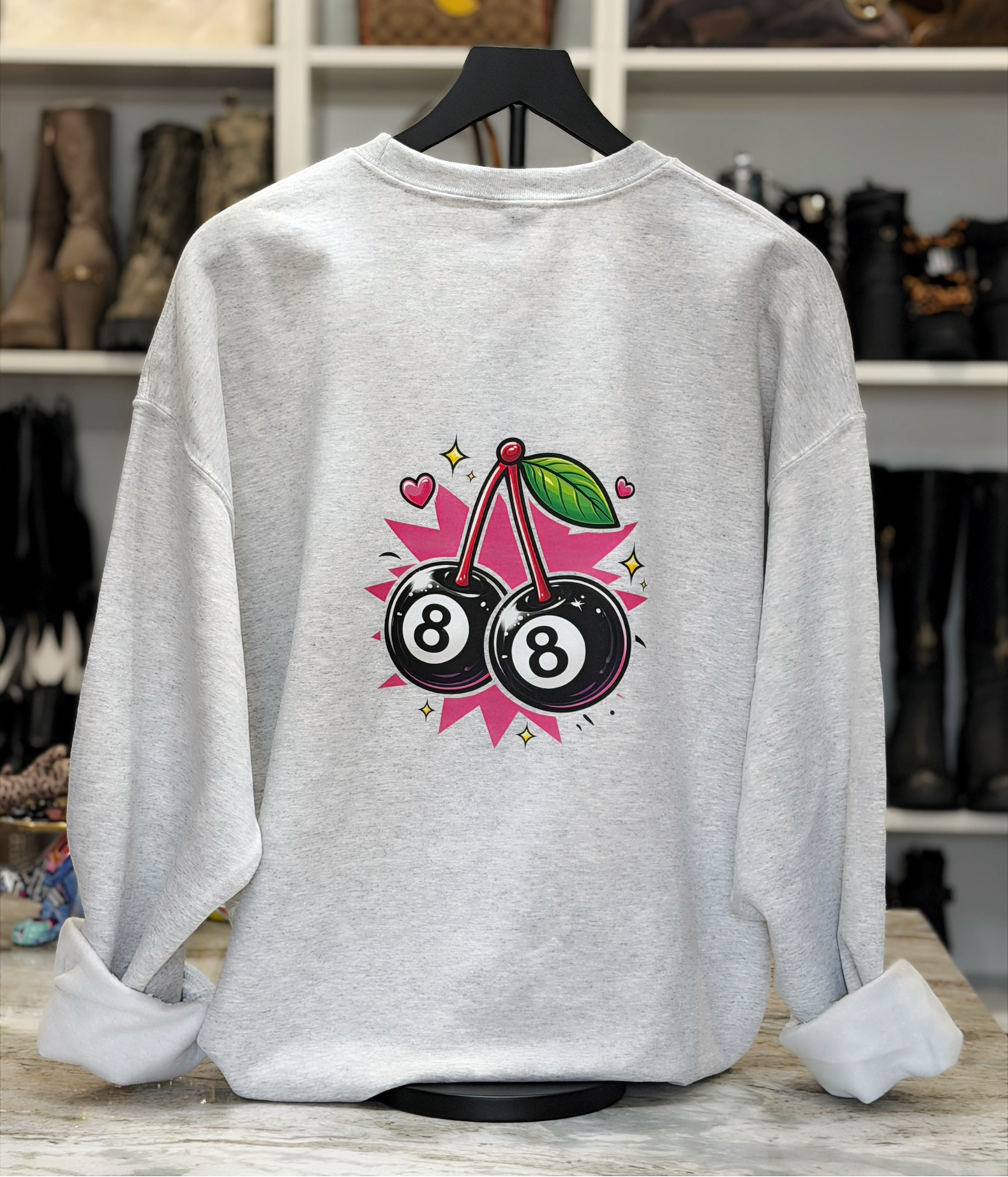 Gray sweatshirt with 8-ball and cherry design on a hanger against a shoe store background