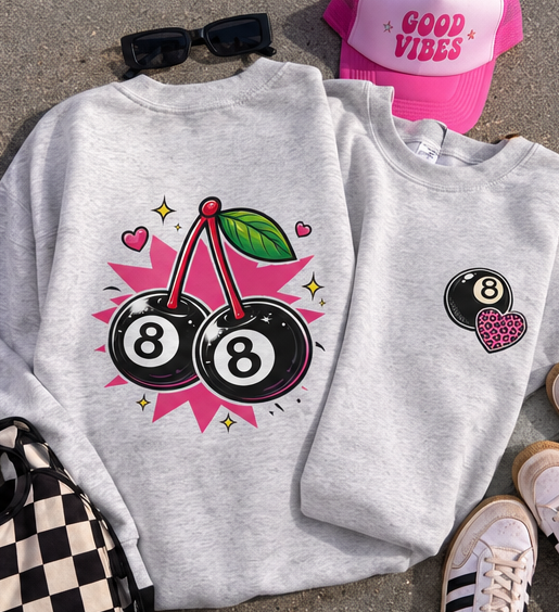 A light heather gray crewneck sweatshirt laid flat on a gray surface, featuring a graphic of two black 8-ball cherries with pink starburst and heart accents on the front. A matching heather gray sweatshirt is folded behind it showing a smaller version of the 8-ball cherry graphic on the left chest.