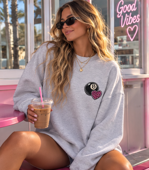 A woman wearing an oversized light gray sweatshirt with a black and pink 8-ball and heart graphic on the chest, sitting on a pink bench and holding an iced coffee.