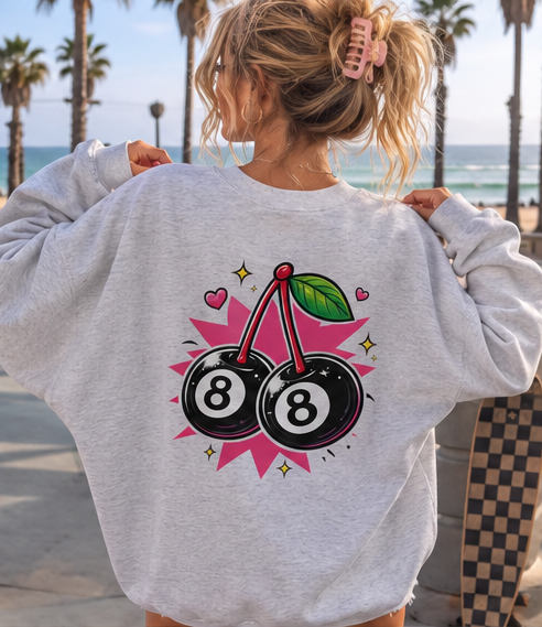 A woman viewed from behind wearing a light heather gray sweatshirt with a graphic of two 8-ball pool balls connected by stems, surrounded by pink hearts and stars. The background shows a beach boardwalk with palm trees and the ocean.