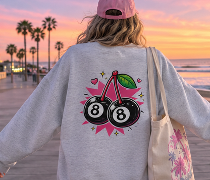 A person standing on a boardwalk at sunset wearing a light gray oversized sweatshirt with a large graphic of two 8-balls and pink hearts on the back, a pink cap, and carrying a floral tote bag.