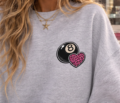 A close-up of a person wearing a heather gray crewneck sweatshirt with a graphic of a black 8-ball and a pink leopard-print heart on the chest.