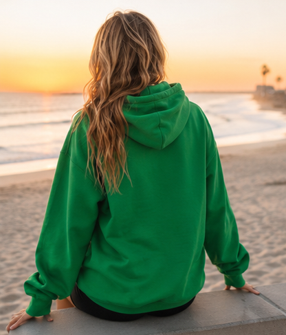 A woman with long wavy light brown hair sits on a concrete ledge at a beach, viewed from behind. She is wearing an oversized bright green hoodie and black pants, with the ocean and a warm sunset sky in the background.