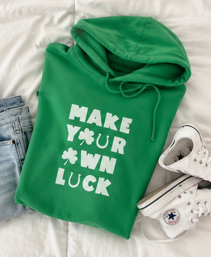 A bright green hoodie with white text reading 'MAKE YOUR OWN LUCK' and a shamrock graphic, styled in a flat lay on white bedding with folded blue jeans and white sneakers nearby.