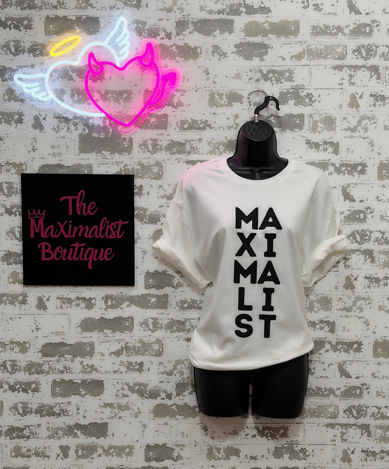 White t-shirt with 'MAXIMALIST' text, neon sign, and 'The Maximalist Boutique' sign on a textured wall.