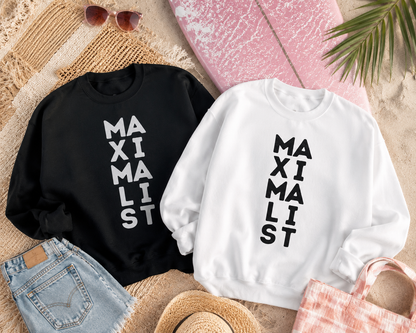 Two crewneck sweatshirts laid flat on a beach-themed background. One sweatshirt is black with the word 'MAXIMALIST' printed vertically in white, and the other is white with the same text printed vertically in black.